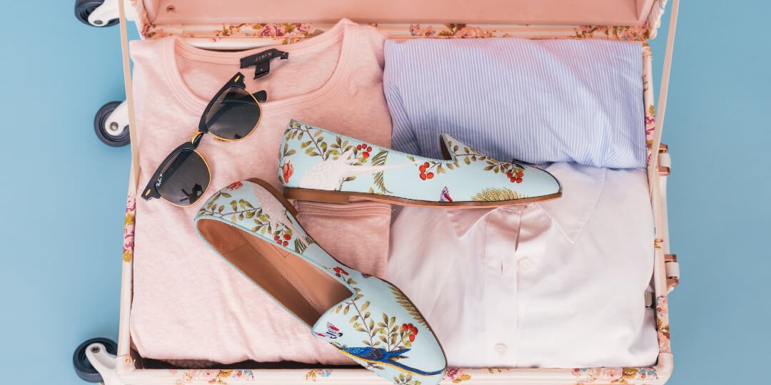A light pink suitcase holds pink tops, a blue top, sunglasses, and blue floral patterned loafers on a light blue background.