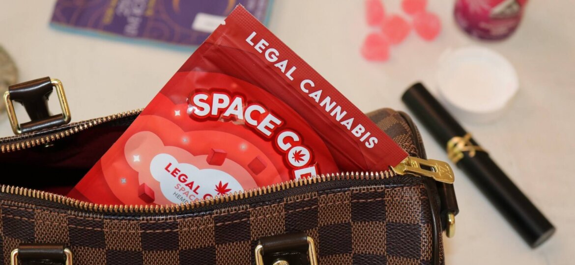 A purse is open with legal cannabis inside. On the table below are cannabis products such as a vape and gummies.